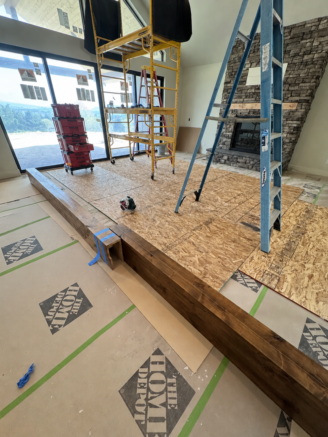 Wood beam assembly in a living room during installation, with stone fireplace, scaffold and ladder.