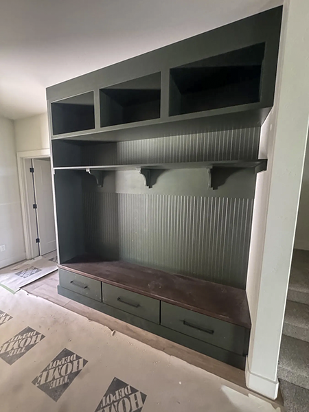 Deep-green built-in entry bench with beadboard backing, open cubbies, a wood seat and three drawers, with floor protection still in place.