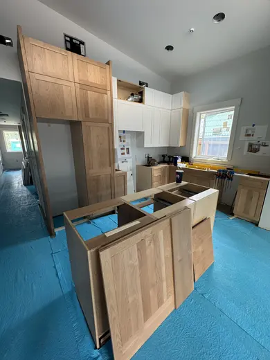 Natural wood kitchen cabinets and an island during installation over blue floor protection, view 2.